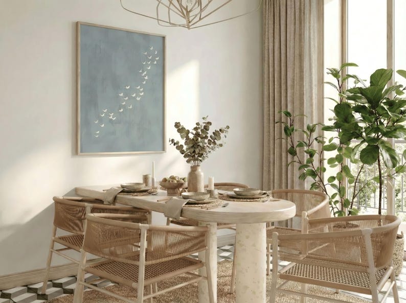 May include: A dining room scene featuring a round table with woven chairs. A framed artwork with a blue background and white bird silhouettes hangs on the wall. A vase of dried flowers and place settings adorn the table. Natural light streams in from a large window.