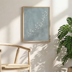 May include: A framed art print featuring a flock of white birds in flight against a muted blue-grey background. The artwork is displayed in a light wood frame. A wooden chair and a potted plant with large green leaves are in the foreground.