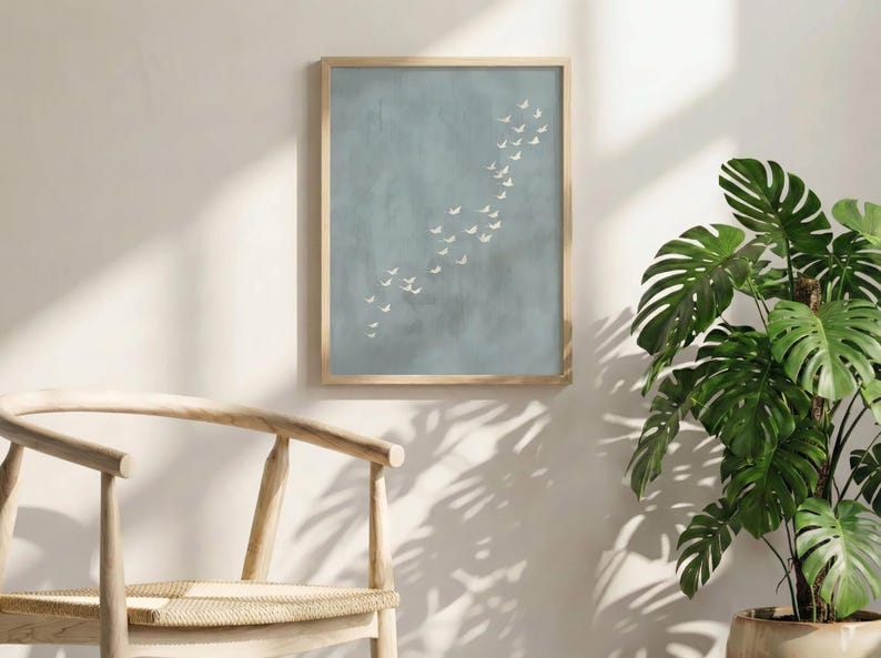 May include: A framed art print featuring a flock of white birds in flight against a muted blue-grey background. The artwork is displayed in a light wood frame. A wooden chair and a potted plant with large green leaves are in the foreground.