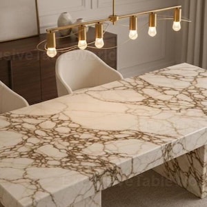 Puede incluir: Una mesa de comedor rectangular de mármol con un patrón veteado marrón, sostenida por una base a juego. La mesa está rodeada por cuatro sillas de color crema. Una lámpara dorada cuelga sobre la mesa.