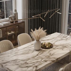 May include: A marble-topped dining table with a rectangular shape, surrounded by cream-colored chairs. The table is set with a vase of dried flowers and a plate of fruit. A wooden cabinet and decorative items are visible in the background.