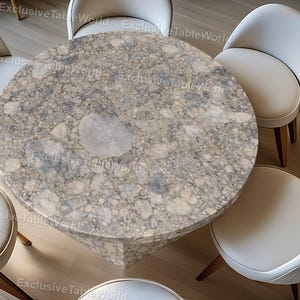 May include: A round table with a gray and white speckled surface, surrounded by six white chairs with wooden legs. The table's base is a solid, geometric shape. The chairs have curved backs and light-colored upholstery. The table is approximately 122 cm in diameter.