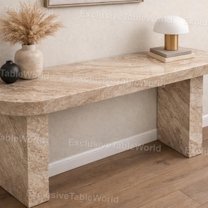 Puede incluir: Una consola de mármol beige con una parte superior redondeada y dos patas rectangulares. La mesa está decorada con un jarrón de flores secas, una esfera negra y una pequeña lámpara blanca y dorada. La mesa está sobre un suelo de madera.