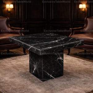 May include: A square black marble coffee table with white veining. The table has a solid, rectangular base and a matching top. The table is in a room with brown leather chairs and a neutral-coloured rug. The text "Exclusive TableWorld" is visible.