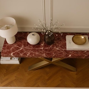 Puede incluir: Mesa de centro moderna con tapa de mármol rojo y patas cruzadas doradas. Decorada con una lámpara blanca, un jarrón blanco, un jarrón oscuro con flores, libros y un cuenco dorado. La mesa mide 120 cm de largo.