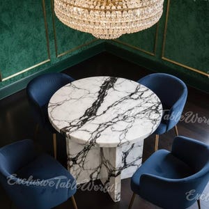 May include: A circular marble table with black veining, encircled by four blue velvet chairs. A large crystal chandelier hangs overhead. The table has a pedestal base. The room features green walls and dark wood flooring.