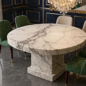May include: A circular marble dining table with a white and grey veined pattern. The table is supported by a square pedestal base. Green velvet chairs with wooden legs surround the table, creating an elegant dining set.