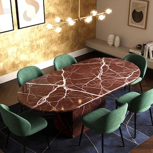 Puede incluir: Un comedor moderno con una gran mesa ovalada. La mesa tiene una tapa de mármol rojo con vetas blancas. Seis sillas de terciopelo verde rodean la mesa. Una lámpara de araña dorada cuelga encima.