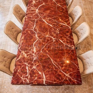 Puede incluir: Una mesa de comedor rectangular con tapa de mármol rojo y vetas blancas. Ocho sillas beige tapizadas rodean la mesa. La mesa mide aproximadamente 3 metros de largo.