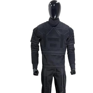Op de afbeelding: Zwart tactisch uniform op een mannequin. Het uniform bestaat uit een top met lange mouwen en verstevigde panelen en een hoge kraag, samen met een bijpassende broek. Witte stiksels omlijnen het ontwerp en de top heeft verschillende klittenbandpatches.