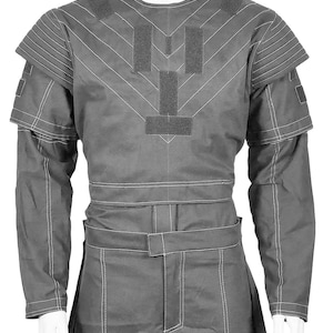 May include: A gray, long-sleeved garment with white stitching and a structured design. The top features shoulder pads and a series of rectangular patches. The garment is displayed on a mannequin.