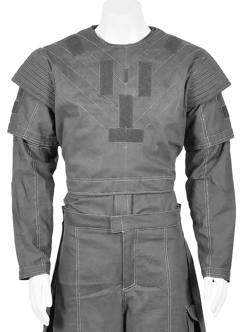 May include: A gray, long-sleeved garment with white stitching and a structured design. The top features shoulder pads and a series of rectangular patches. The garment is displayed on a mannequin.
