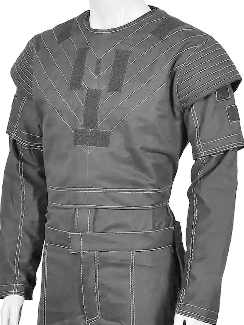 May include: A gray protective suit with white stitching and multiple Velcro patches. The suit features a round neck, long sleeves, and a belt. The shoulders have padded sections, and the suit appears to be made of a durable material.