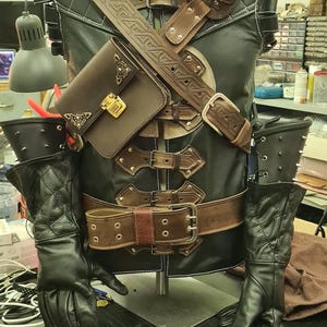 May include: A black leather costume with brown detailing, including a vest, belt, and shoulder straps. The costume features intricate stitching and detailing, with a small brown satchel and black gloves. The costume is displayed on a mannequin.