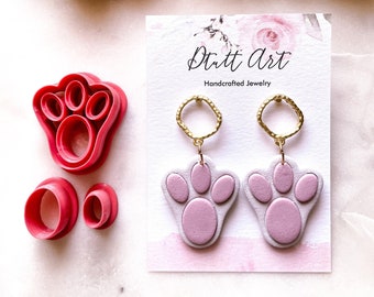 May include: A pair of purple paw print earrings with gold hoops. The earrings are handmade and are displayed on a white card with the text "Dutt Art Handcrafted Jewelry".