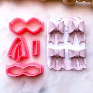 May include: Pink polymer clay cutters shaped like a bow, a flower, and a rectangle. Four purple polymer clay bows are displayed on a white card with the text "tied with love".