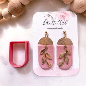 Könnte beinhalten: Ein Paar rosa und goldene Ohrringe mit einem Blattdesign. Die Ohrringe sind handgefertigt und werden auf einer weißen Karte mit dem Text "Dutt Art Handgefertigte Schmuckstücke" präsentiert.