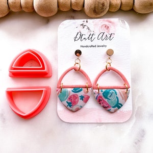 Puede incluir: Un par de pendientes geométricos rosa y blanco con estampado de sandía y fruta. Los pendientes están hechos a mano y presentan detalles dorados. Los pendientes se muestran en una tarjeta blanca con el texto "Dritt Art Joyería artesanal".