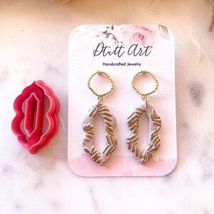 Puede incluir: Un par de pendientes colgantes con una base gris claro y un patrón de hojas doradas. Los pendientes están en una tarjeta blanca con el texto "Stutt Art Joyería artesanal".