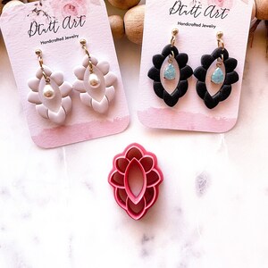 May include: Two pairs of handmade earrings on white cards with the text "Dutt Art Handcrafted Jewelry". The earrings on the left are white with a pearl accent. The earrings on the right are black with a light blue stone accent. A pink plastic mold for making earrings is in the foreground.