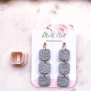 May include: A pair of gray polymer clay earrings with a rose pattern. The earrings are made of three square shapes stacked on top of each other. The earrings are hanging from gold hooks. The earrings are on a white card with the text "Dutt Art Handcrafted Jewelry".