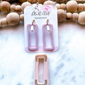 Puede incluir: Un par de pendientes rectangulares de color rosa con una perla colgante. Los pendientes están en una tarjeta blanca con el texto "Dutt Art Joyería artesanal".