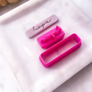 Puede incluir: Tres cortadores de galletas rosas sobre una superficie de mármol blanco. Los cortadores tienen forma de las palabras "esperanza" y "oración" y un rectángulo.