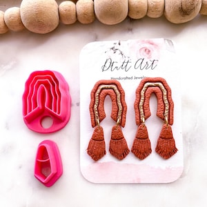 Può includere: Tre paia di orecchini in argilla polimerica color terracotta con dettagli dorati e frange. Gli orecchini sono esposti su una carta bianca con il testo "Dutt Art Handcrafted Jewels".