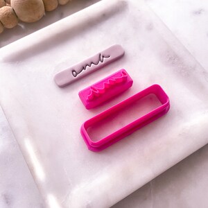 以下が含まれることがあります： ピンクとパープルのクッキーカッター3個セット。カッターは長方形、波線、そして「amber」という単語の形をしています。