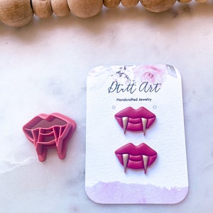 May include: A pair of red and gold vampire fang earrings on a white card with the text "Dutt Art Handcrafted Jewelry".