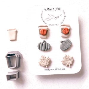 May include: A set of six fall-themed earrings featuring a coffee cup, pumpkin, and leaf designs. The earrings are made of polymer clay and come in shades of brown, orange, and green. The earrings are packaged on a white card with the text "D tutt Art" and "Studs Pack".