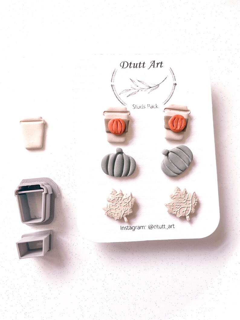 May include: A set of six fall-themed earrings featuring a coffee cup, pumpkin, and leaf designs. The earrings are made of polymer clay and come in shades of brown, orange, and green. The earrings are packaged on a white card with the text "D tutt Art" and "Studs Pack".