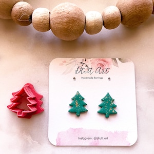 Peut inclure: Une paire de boucles d'oreilles en forme de sapin de Noël vertes avec des paillettes dorées. Les boucles d'oreilles sont faites à la main et sont présentées sur une carte blanche avec le texte "Dutt Art Handmade Earrings" et "Instagram: @dutt_art".