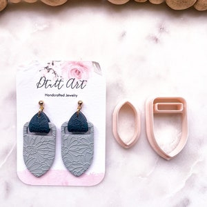 May include: A pair of gray and black floral earrings with gold accents. The earrings are displayed on a white card with the text "Dutt Art Handcrafted Jewelry".