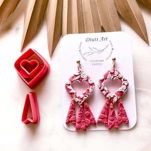 May include: Pink and white heart-shaped dangle earrings with a floral pattern. The earrings are handmade and have a gold-coloured hook.