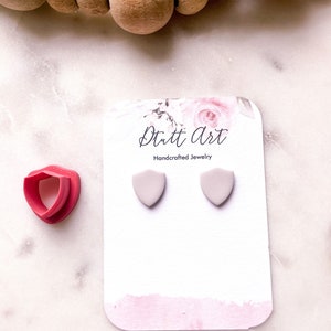 以下が含まれることがあります： 「Ditt Art Handcrafted Jewelry」の文字が入った白いカードに、薄い灰色のシールド型スタッドピアスが2つあります。赤いシールド型の粘土カッターがピアスの隣にあります。背景には木製のビーズがあります。