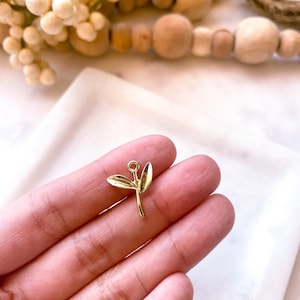May include: A gold-colored leaf charm with two leaves and a small loop at the top. The charm is held in a hand against a white background. The charm is likely for jewelry making or crafting.
