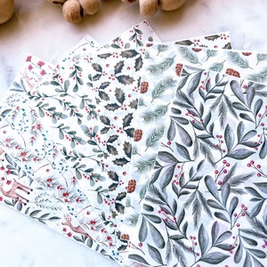 Puede incluir: Cinco hojas de papel de regalo diferentes con un tema invernal. Los patrones incluyen acebo, ramas de pino, bayas y renos. El papel es blanco con acentos verdes, rojos y marrones.