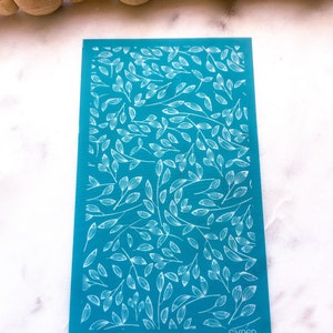 May include: A teal blue nail art stamping plate with a white design of leaves and branches. The plate has the number SY069 printed in the bottom right corner.
