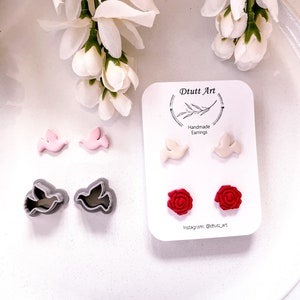 May include: A set of handmade earrings featuring a dove design in white, pink, and gray, and a rose design in red. The earrings are displayed on a white card with the text "D tutt Art Handmade Earrings Instagram: @dtutt_art".