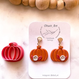 May include: A pair of orange pumpkin earrings with white flowers and green leaves. The earrings are handmade and have gold hooks. The earrings are displayed on a white card with the text "D tutt Art" and "Handmade Earrings".