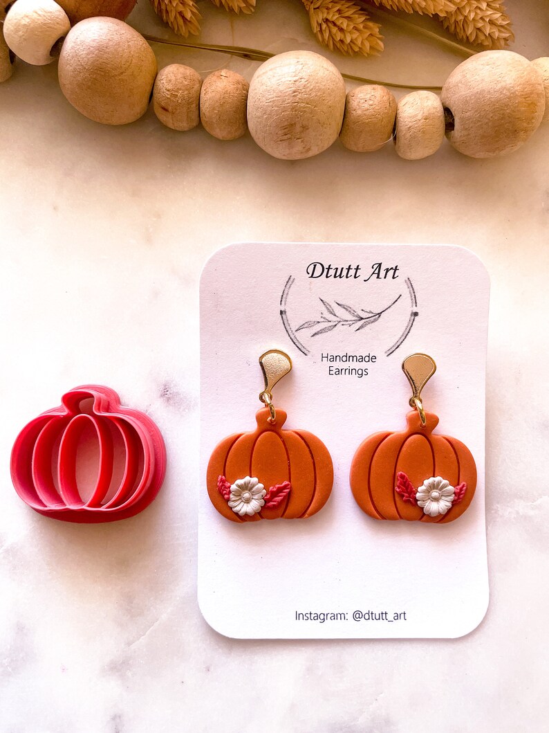May include: A pair of orange pumpkin earrings with white flowers and green leaves. The earrings are handmade and have gold hooks. The earrings are displayed on a white card with the text "D tutt Art" and "Handmade Earrings".