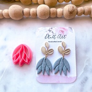 May include: A pair of grey polymer clay earrings with gold leaf accents. The earrings are shaped like a flower with five petals. The earrings are on a white card with the text "Dutt Art Handcrafted Jewelry".