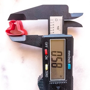 May include: A black digital caliper measuring a red plastic cookie cutter in the shape of a heart. The caliper reads 0.58 inches.