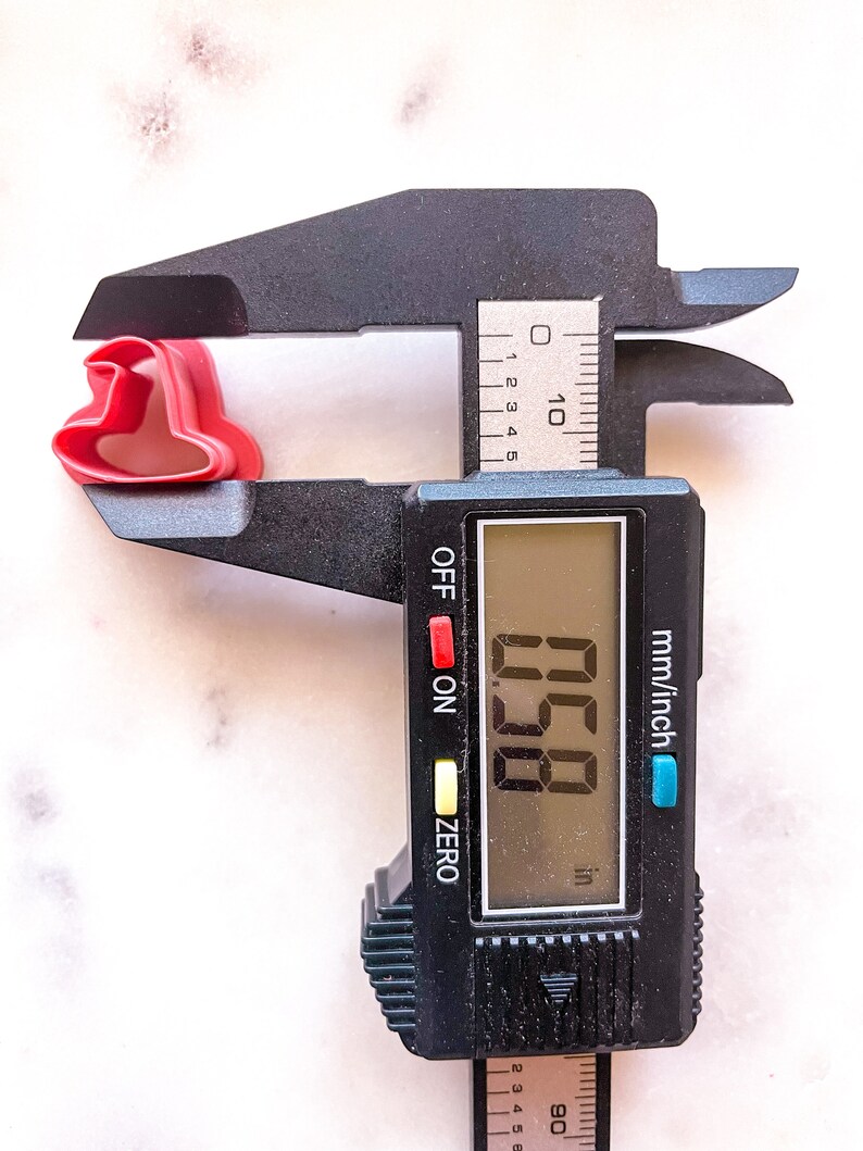 May include: A black digital caliper measuring a red plastic cookie cutter in the shape of a heart. The caliper reads 0.58 inches.