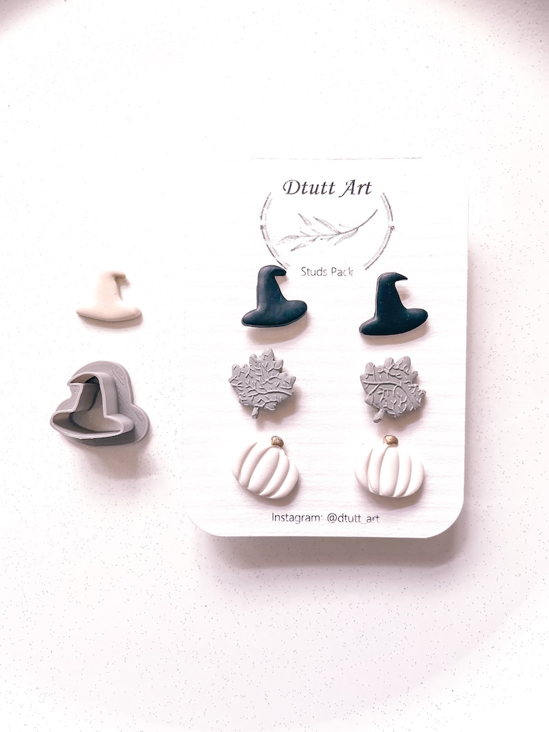 May include: A set of six Halloween-themed earrings, including witch hats, pumpkins, and leaves. The earrings are made of polymer clay and come in black, white, and gray colors. The card reads "Dtuutt Art" and "Studs Pack".