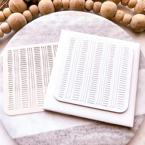 May include: Two white, square, textured stencils with a repeating pattern of vertical lines. The stencils are on a white marble surface.