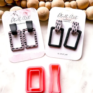 Puede incluir: Dos pares de pendientes hechos a mano con marcos negros y un diseño estampado en rosa y negro. Los pendientes se muestran en tarjetas blancas con el texto "Dutt Art Handmade Earrings" y "IG: DTUTT_ART". También se muestran dos cortadores de galletas de plástico rosa en la imagen.