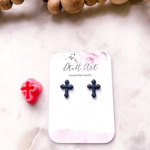 Puede incluir: Un par de pendientes de cruz negros en forma de clavo sobre una tarjeta blanca con el texto "Dutt Art Joyería artesanal".