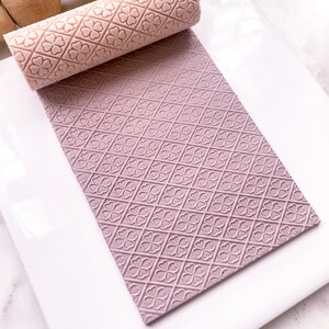May include: A pink rolling pin with a clover pattern is shown on a white surface. The rolling pin is used to create an embossed pattern on a sheet of pink fondant.